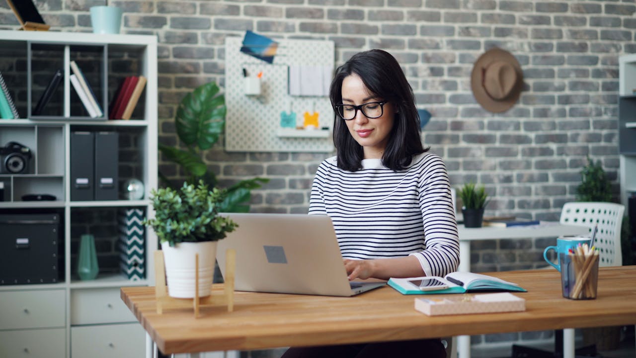 Young woman working on a laptop in a modern, stylish office setting with brick wall decor.
