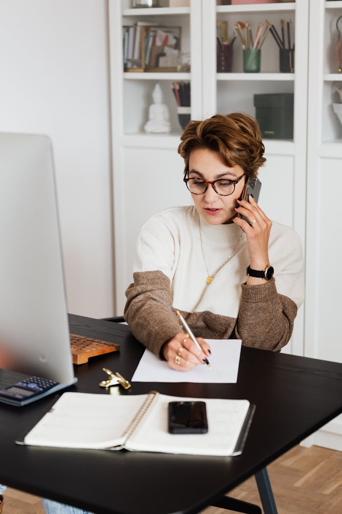 Female remote worker listening attentively and noting down details of project while having phone conversation with director in cozy home study