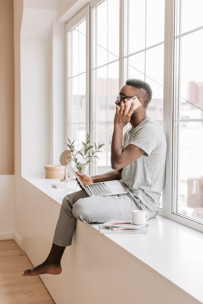 A man sitting by a window, working from home with a laptop and phone, multitasking efficiently.