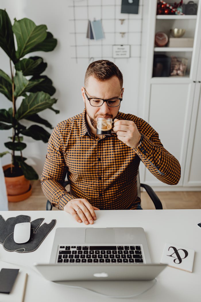 project-01-d Bearded man in glasses drinks espresso while working on a laptop in a home office with plants.