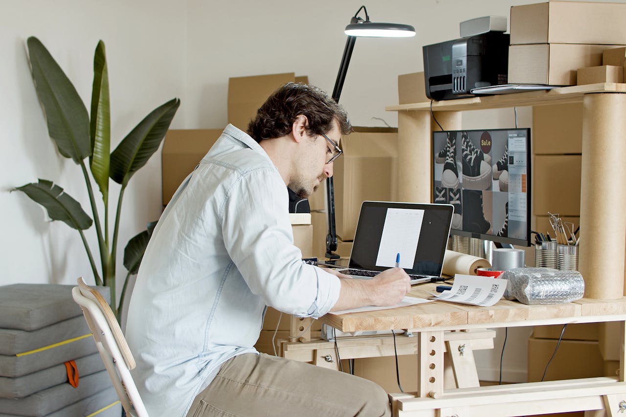 project-02-a Businessman managing an online store in a home office setting, typing on a laptop with packaging supplies around.