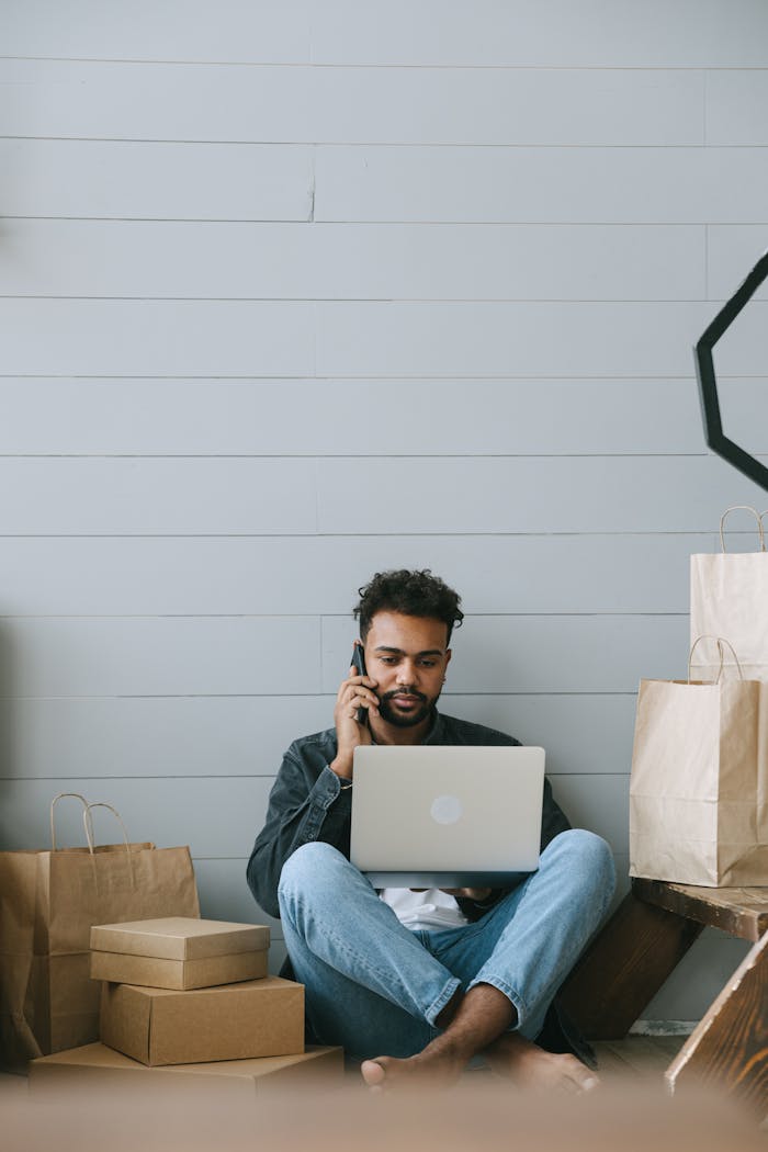 A man engaged in remote work sitting with laptop and phone surrounded by paper bags and boxes.