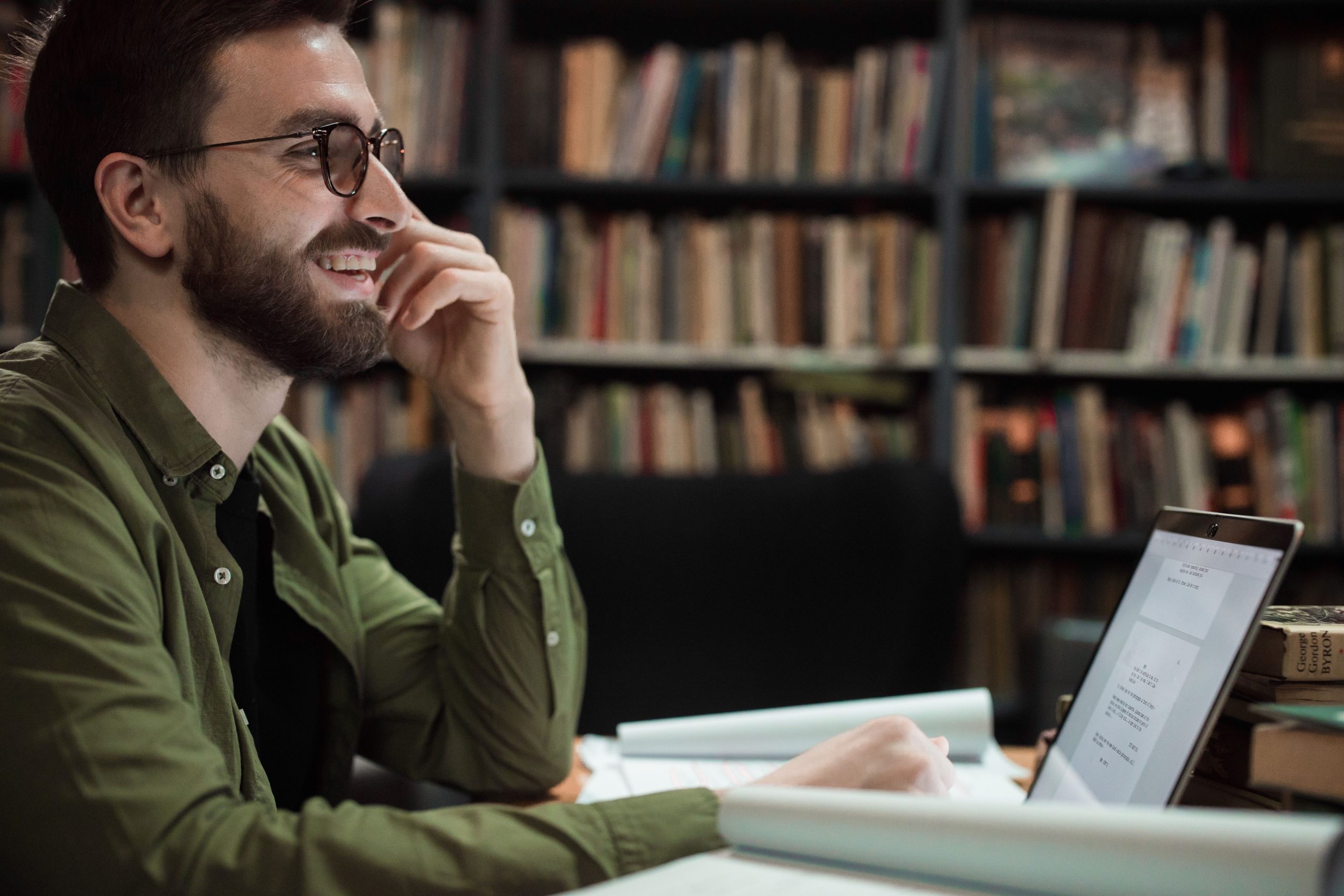 A bearded man in glasses smiles while working on a laptop in a library.